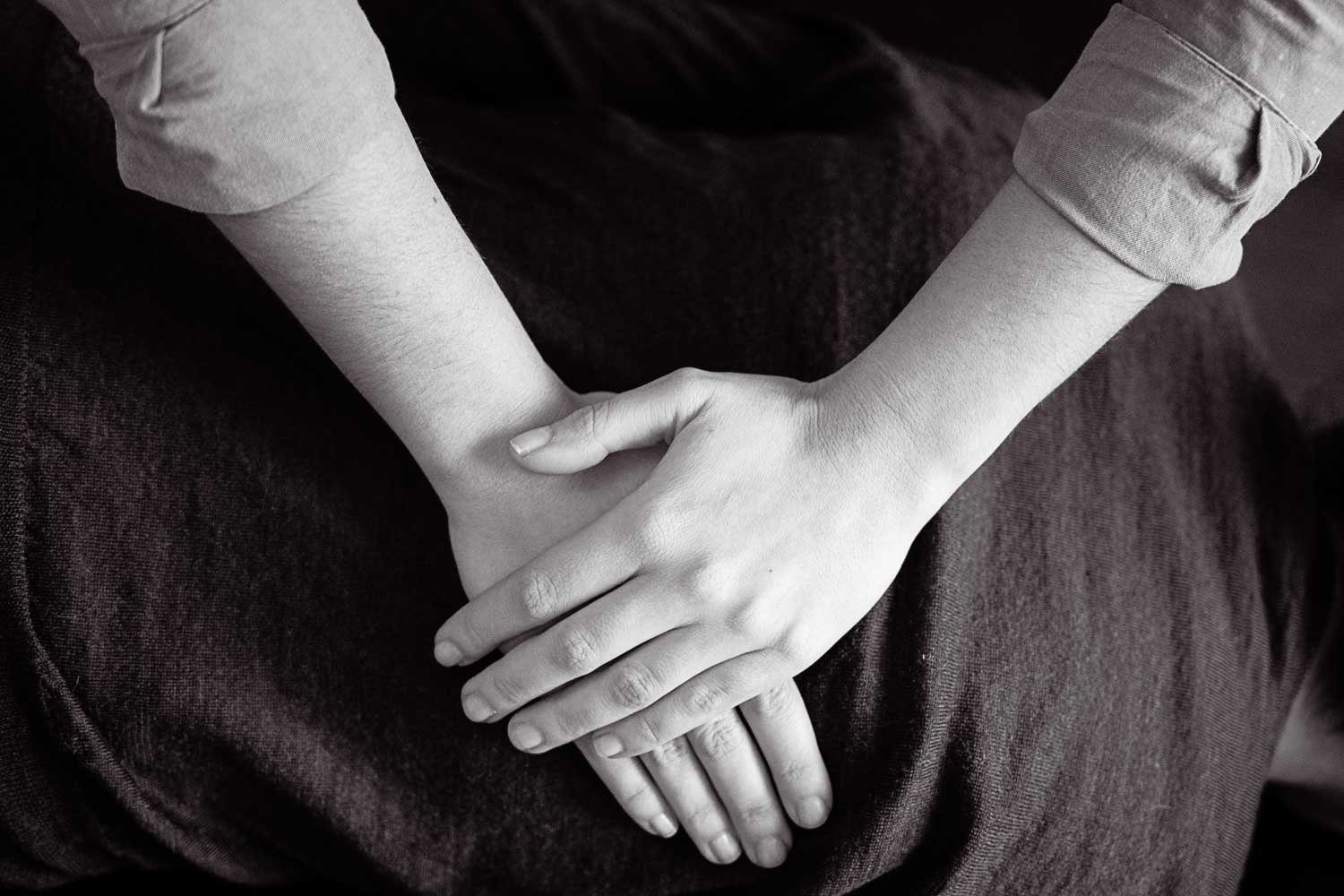 Gros plan noir et blanc des mains de Claire croisées au repos sur un pantalon de lin sombre